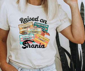On this shirt, there are 5 cassette tapes that belong to the one and only Shania Twain. On these cassette tapes are a few of her songs. They are "Man! I feel like a woman", "Any man of mine", "I'm gonna gotcha good!", "That don't impress me much", and "You're still the one".